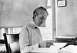 Woman sitting at desk writing, with short hair, long-sleeved white blouse and vest